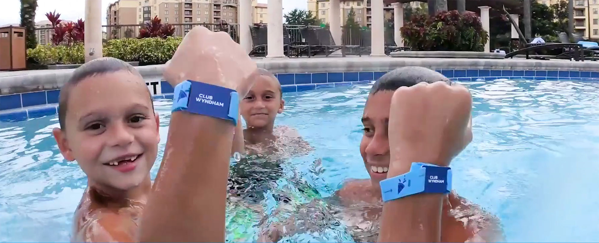 Three boys swimming in a Club Wyndham resort pool wearing Club Wyndham Wristbands on their raised wrists 