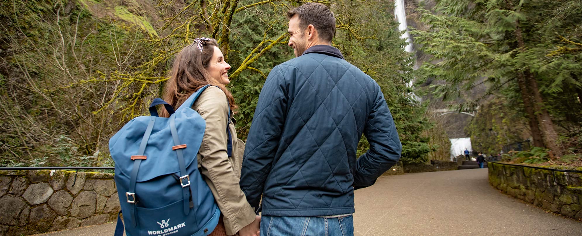 Man and woman holding hands smile and walk towards a waterfall, the woman is wearing a WorldMark by Wyndham backpack.