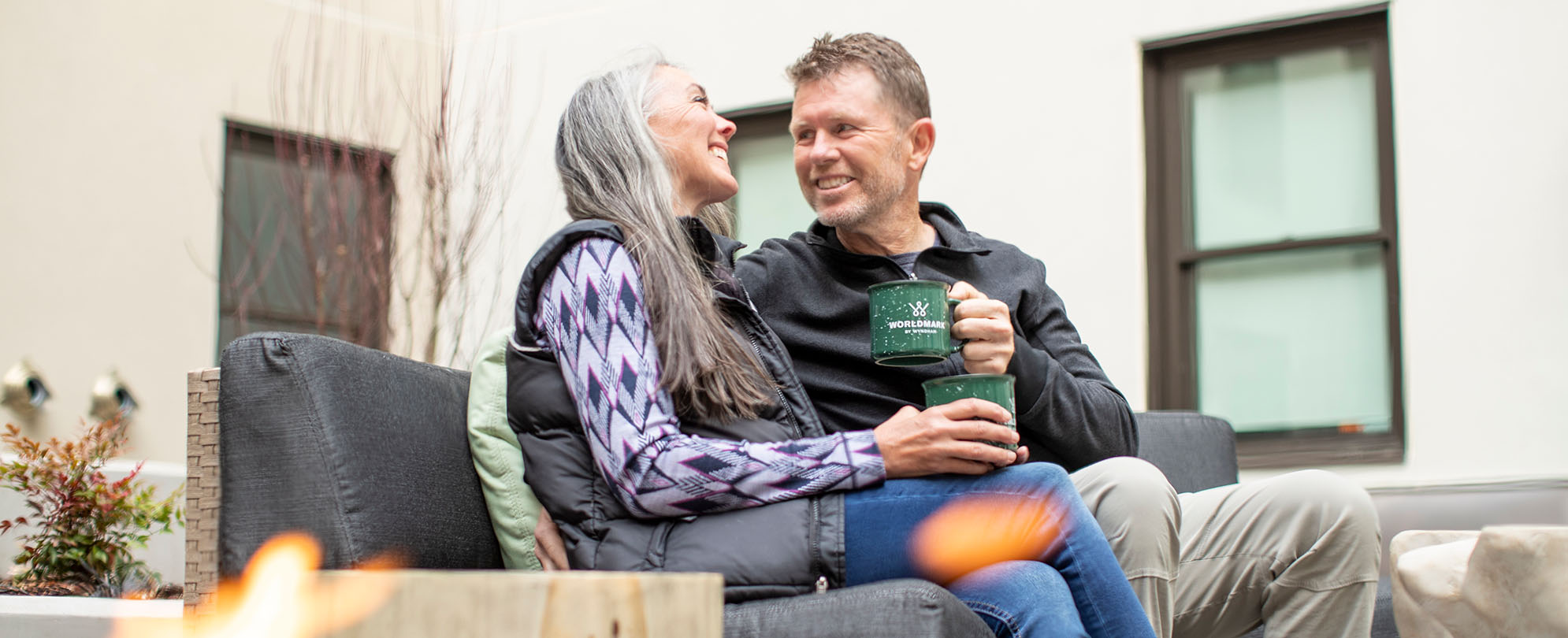 A man and woman holding WorldMark coffee mugs sit on an outdoor couch smiling at each other.